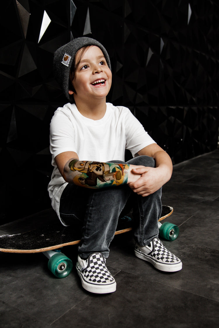 kid sits on skateboard showing off his temporary tattoo on the lower arm. The tattoo shows a robot on a skateboard.